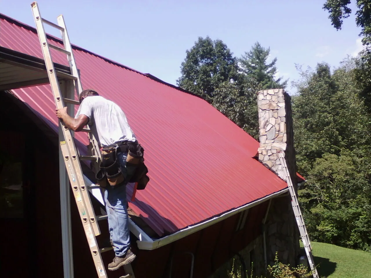 Roofer on a ladder beside a red metal roof and stone chimney — Soffit & Fascia Repair by Roofing Brothers in Perkasie